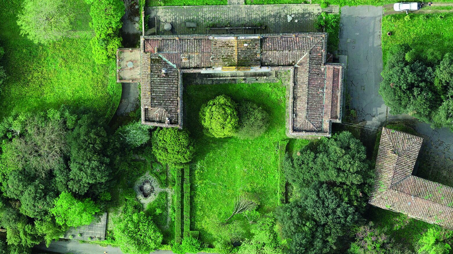 A drone photograph of the villa today, seen from directly above: the horseshoe roof weathered and moss-grown, the courtyard and gardens overgrown, the old parterre traced faintly in the grass.